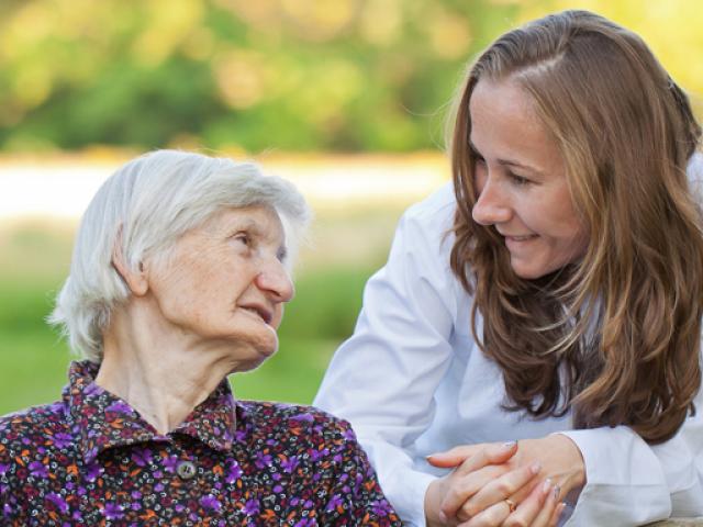An older woman sitting on a parkbench leans back and turns her head to speak with a younger woman who is leaning forward, resting her arms on the back of the parkbench, and smiling.